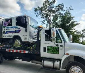 A tow truck transporting a smaller delivery truck with the logo "NOVA AMERICA" on its side. The tow truck is parked on a paved surface, and trees are visible in the background under a partly cloudy sky.