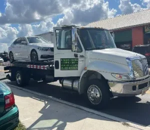 A white tow truck is parked outdoors, carrying a white car on its flatbed. The background shows a building and a partly cloudy sky.