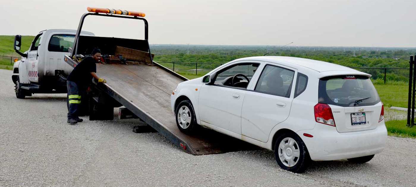 A white car is being loaded onto a flatbed tow truck by a person wearing a black outfit and reflective yellow stripes. The background features grassy fields and a cloudy sky.