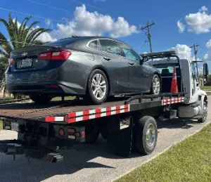 A gray sedan is loaded onto a flatbed tow truck on a sunny day. The truck is parked on the side of the road, with palm trees and power lines visible in the background under a blue sky with clouds.