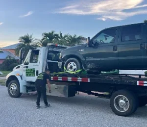 A tow truck is parked on a residential street, with a black van secured on its flatbed. A person is standing near the truck, adjusting equipment. Palm trees and houses are visible in the background, under a partly cloudy sky.