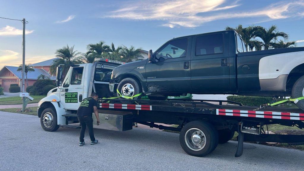 A tow truck is parked on a residential street, loading a dark green pickup truck onto its flatbed. A person stands behind the tow truck, ensuring the vehicle is secured. Palm trees and houses are visible in the background, under a partly cloudy sky.