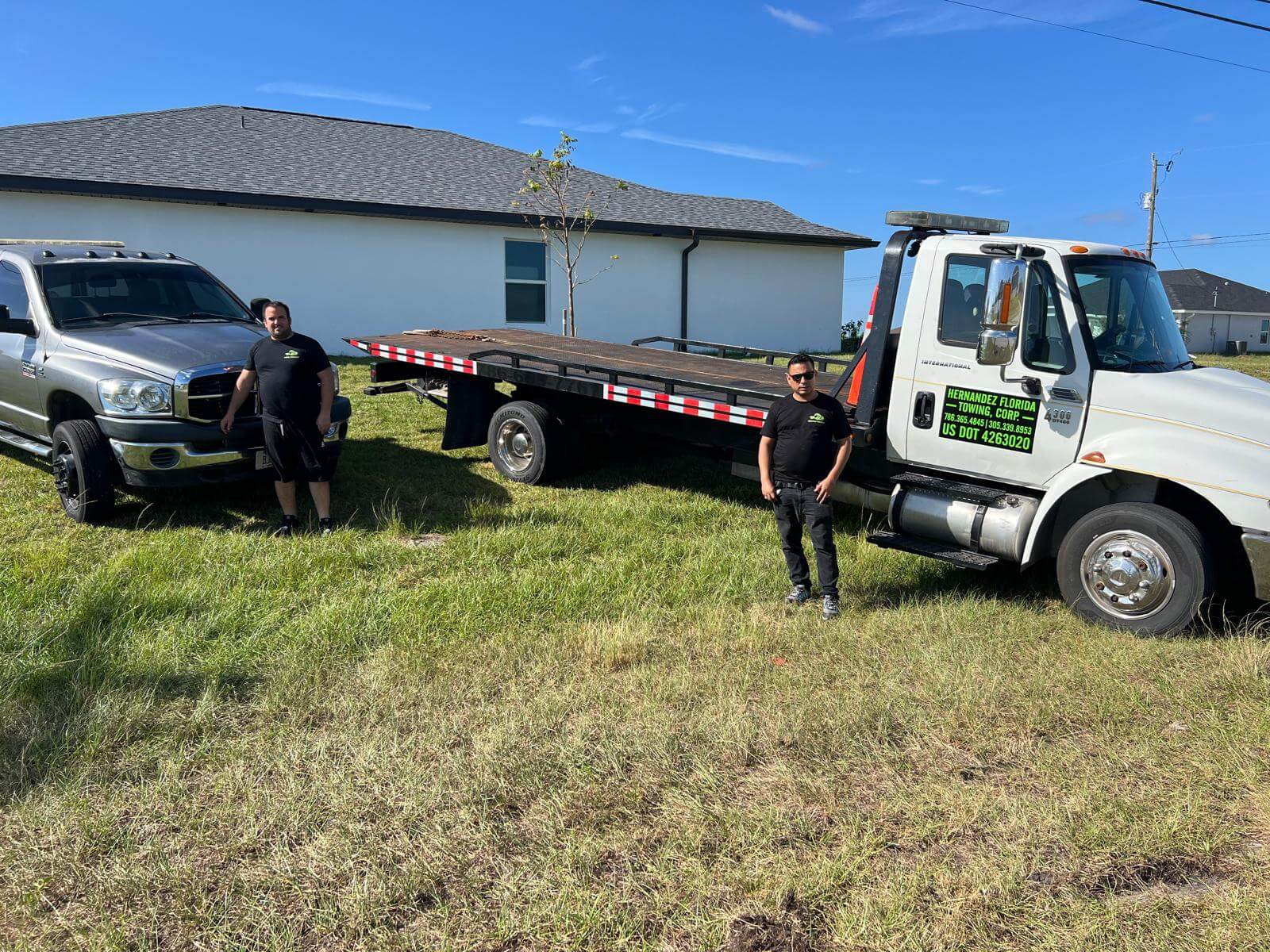 Two people stand in front of a tow truck with a flatbed and a pickup truck on a grassy field. A house is visible in the background, under a clear blue sky.