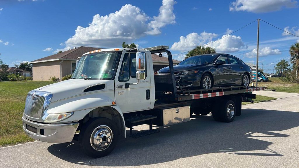 A flatbed tow truck is parked on a sunny residential street, carrying a dark-colored sedan on its back. The sky is blue with scattered clouds, and there are houses and palm trees in the background.