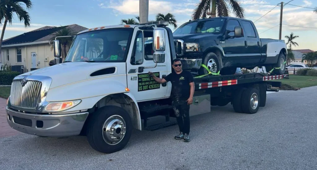 A man stands beside a tow truck on a sunny street. The truck is carrying a black and silver pickup truck on its flatbed. Palm trees and houses are visible in the background. The man is smiling and wearing dark clothing with sunglasses.