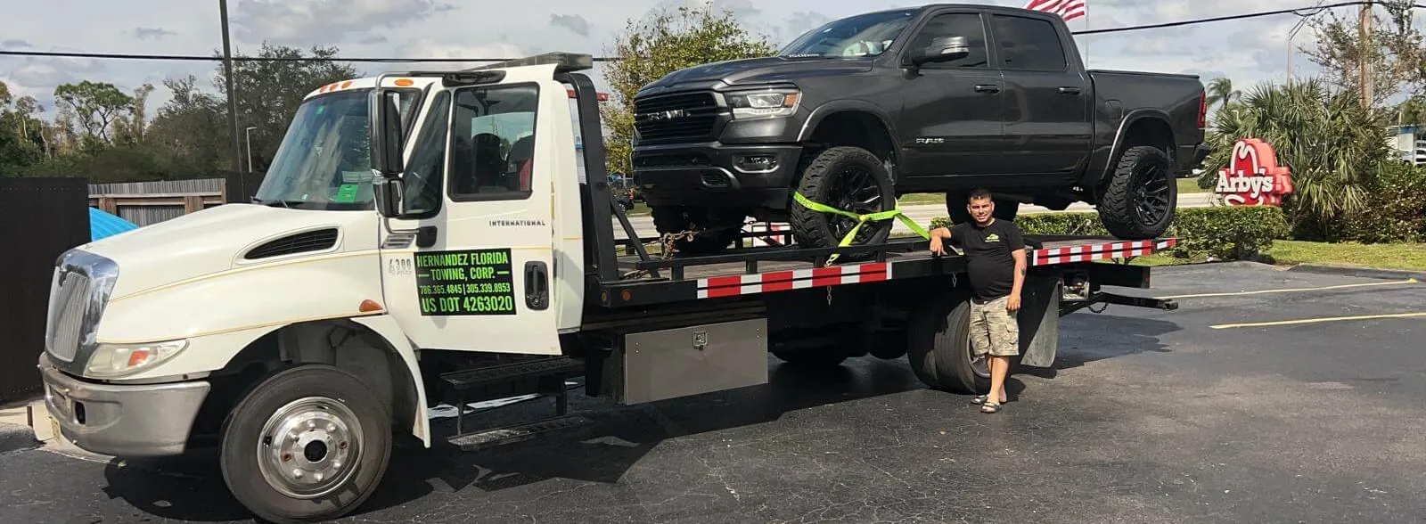 A person stands beside a tow truck in a parking lot. The tow truck is carrying a large black pickup truck secured with green straps. An American flag is visible in the background near some trees.