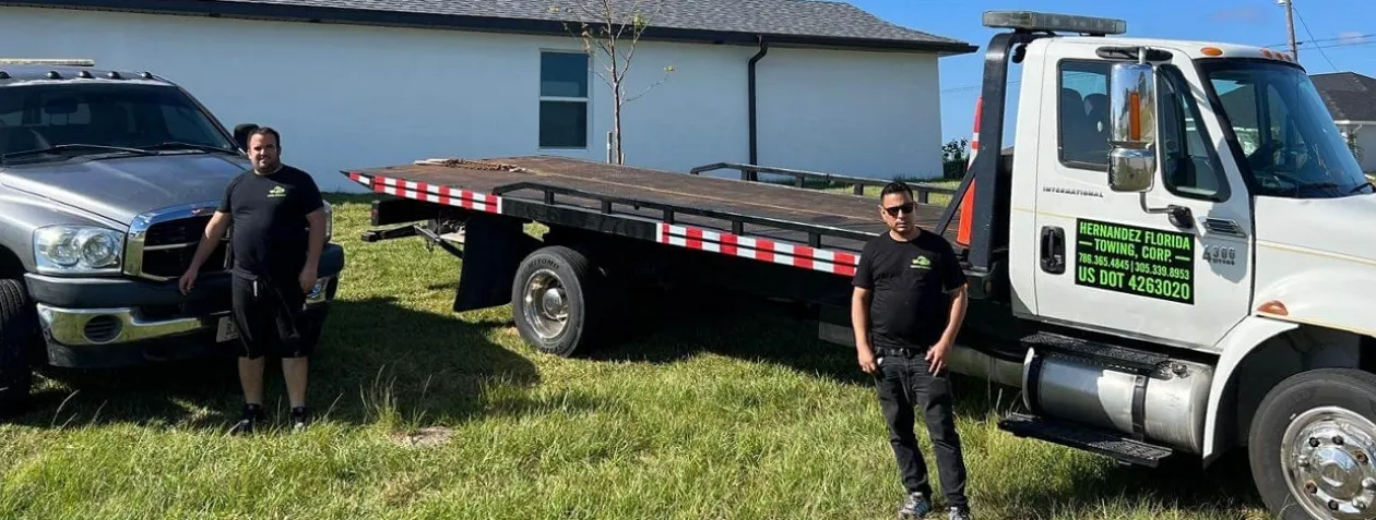 Two men stand beside a silver pickup truck and a white tow truck on a grassy area. The tow truck has a company sign on the door. A white house and clear blue sky are in the background.