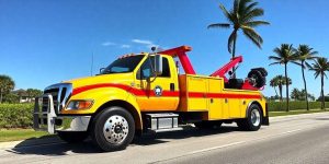 A bright yellow tow truck with red accents is parked on a sunny street. Palm trees and greenery line the road, with a clear blue sky overhead. The truck has a vehicle lift at the back and is positioned in profile view.